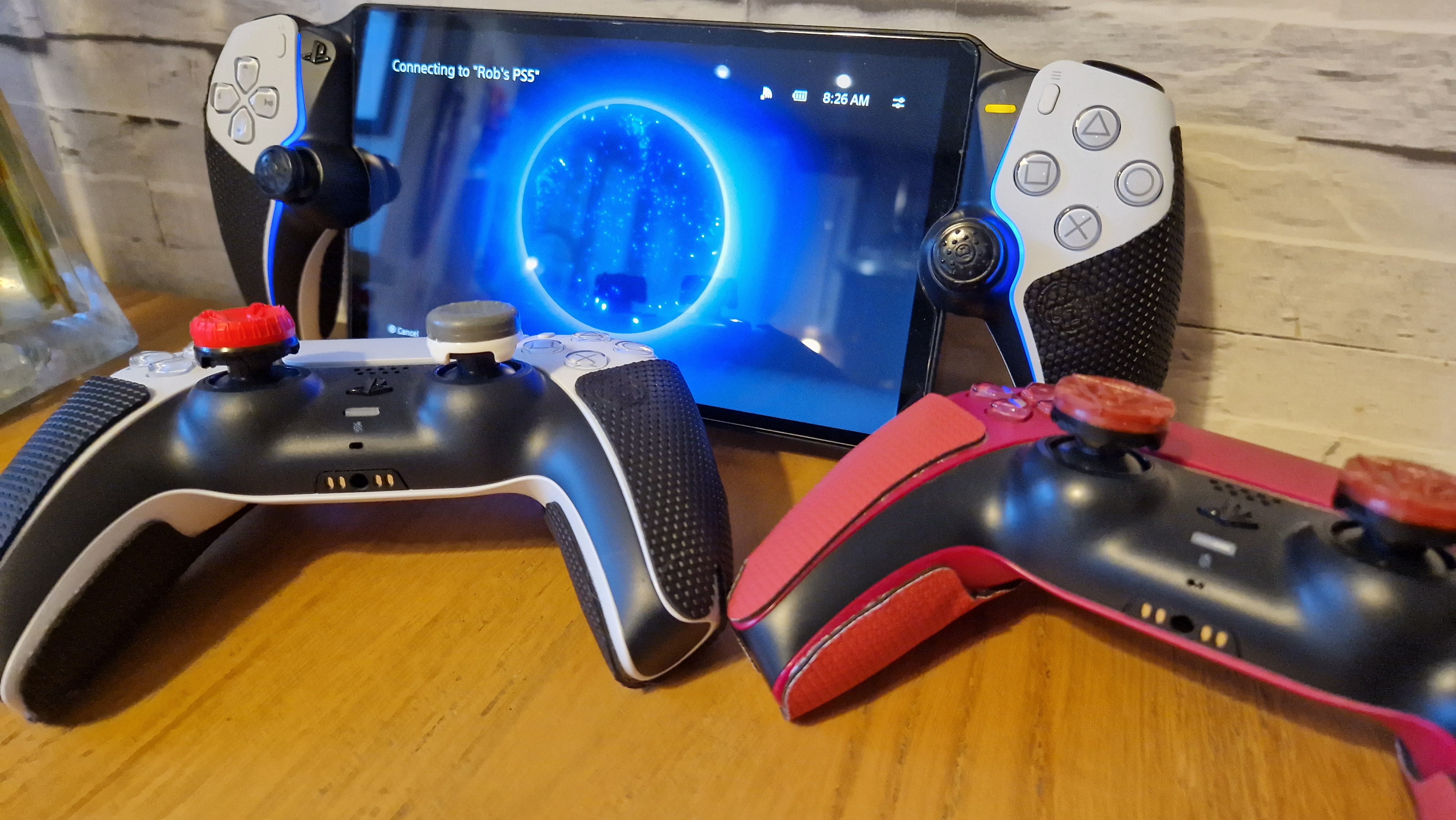 Two DualSense controllers and a PlayStation Portal with thumbsticks and grips attached on a wooden surface with a white brick background