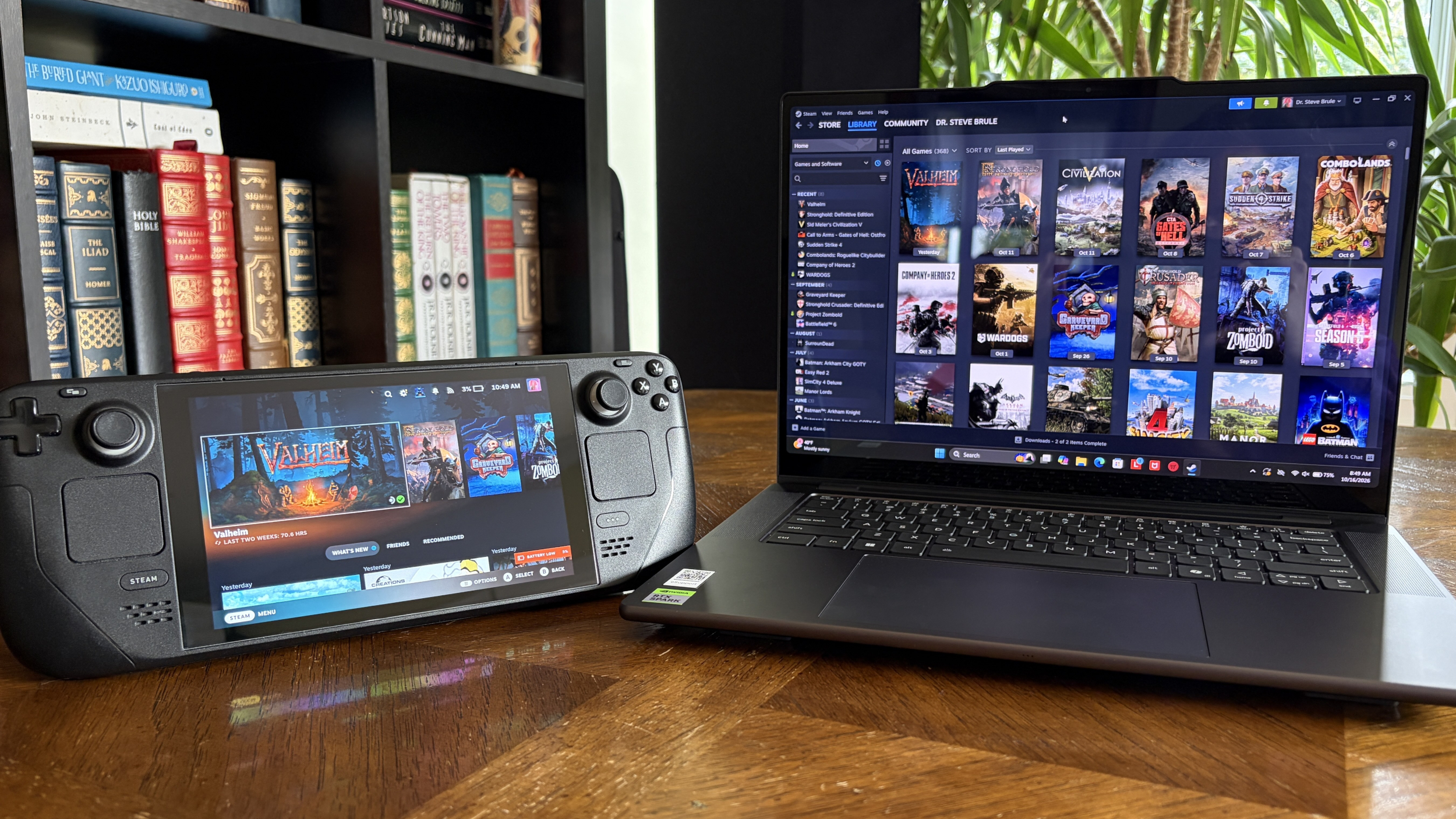 A handheld Steam Deck gaming PC and Lenovo Pro 9n laptop display a dark game library interface on a wooden desk, with books and greenery behind them; cozy, tech-focused mood.
