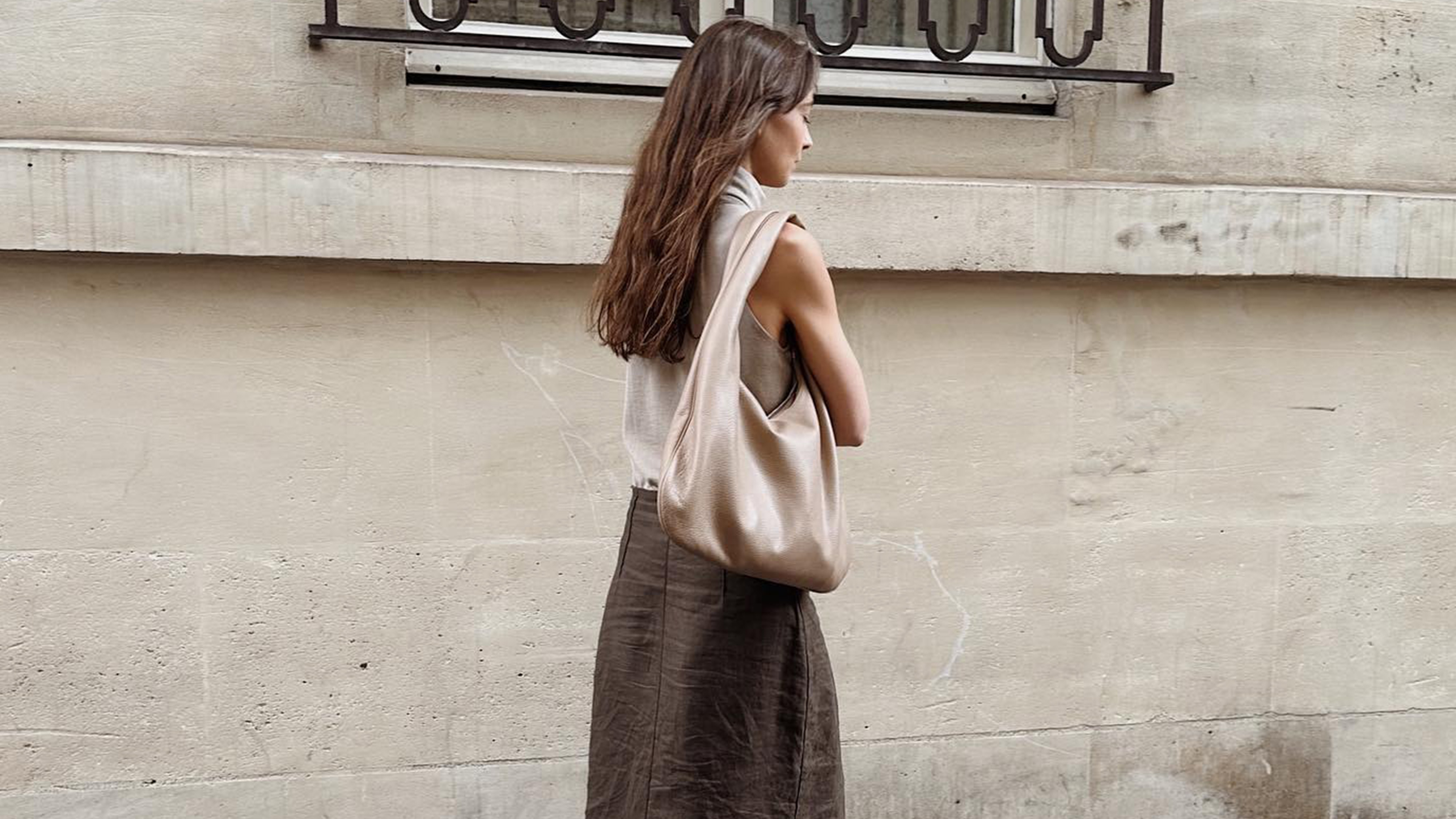 A woman wears a gray top, suede shoulder bag, and brown skirt.