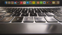 A close-up, low-angle shot shows a MacBook Air keyboard with slightly worn black keys in the foreground, leading up to a blurred macOS dock filled with various application icons on the screen above.