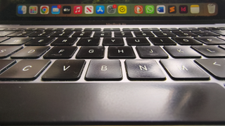 A close-up, low-angle shot shows a MacBook Air keyboard with slightly worn black keys in the foreground, leading up to a blurred macOS dock filled with various application icons on the screen above.