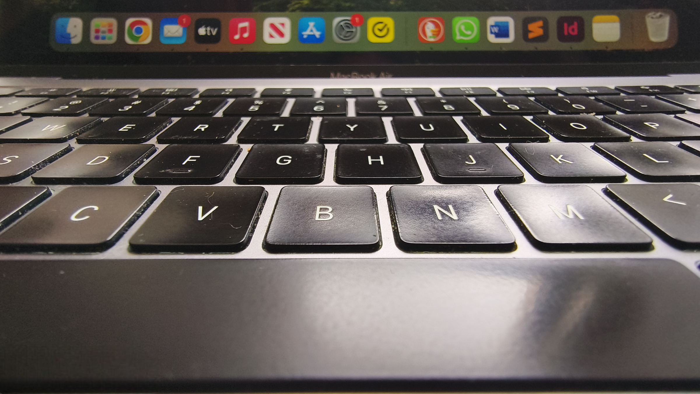 A close-up, low-angle shot shows a MacBook Air keyboard with slightly worn black keys in the foreground, leading up to a blurred macOS dock filled with various application icons on the screen above.