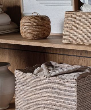 A small round storage basket with a lid on a wooden shelf, surrounded by other woven baskets