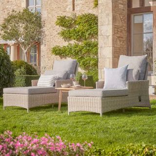 Two wicker sun loungers in a garden with a stone country house in the background
