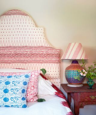 Colorful table lamp with pink striped shade, large rounded headboard in red and white patterned, colorful bedding and cushions, wooden bedside table