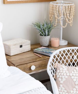 A wooden beaded nightstand with a lamp and notepad on it, next to a white chair with a pillow and a white bed