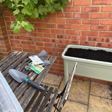 elho Green Basics Grow Table next to wooden garden table and chair, seed packets, garden gloves and GARDENA trowel