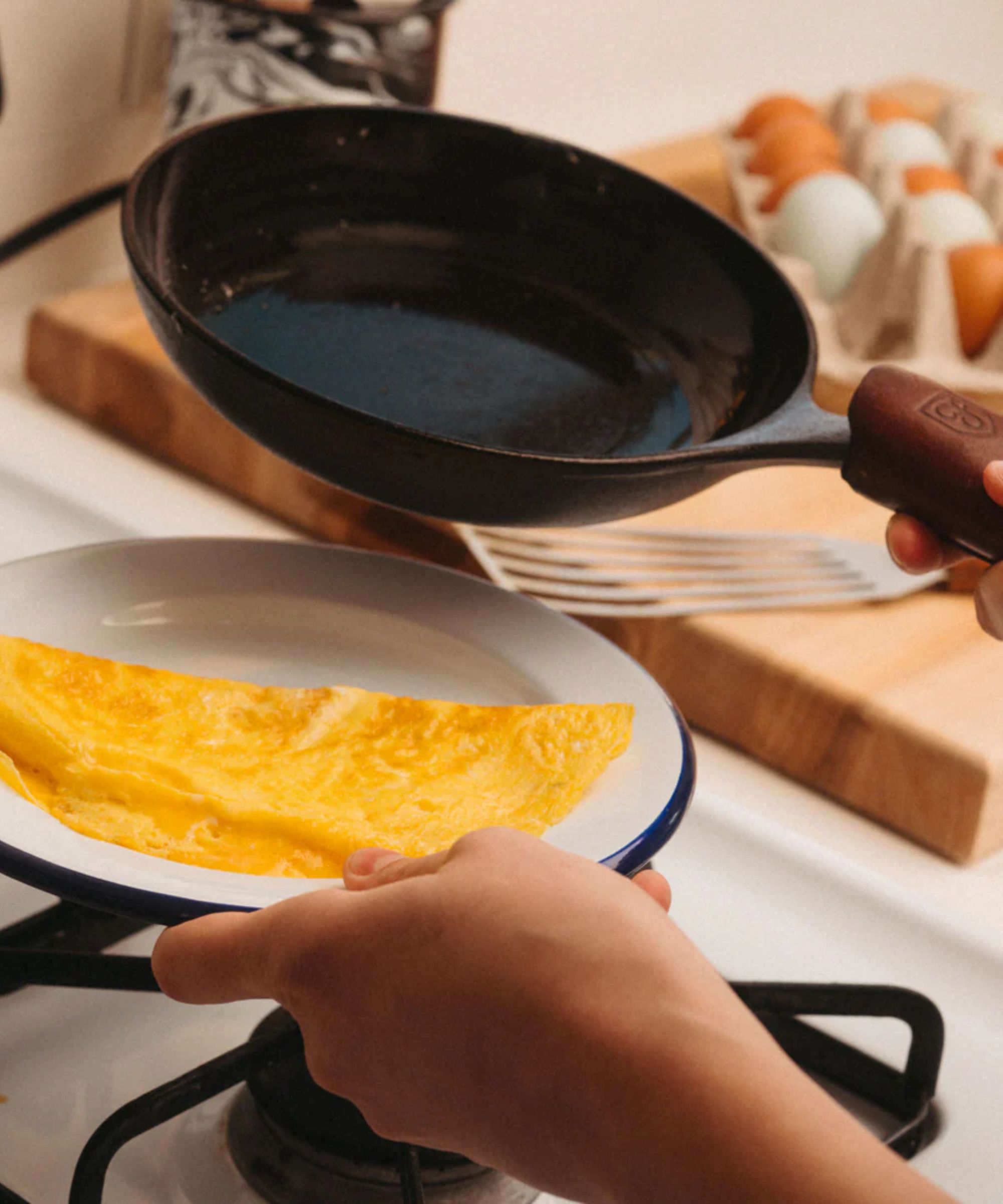Someone holding a small cast iron chef skillet beside a plate with a golden folded omelette, over a stove.