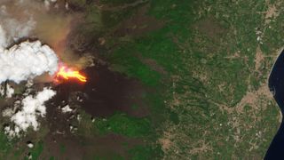 A green land mass with streaks of red and white clouds above it showing a volcano eruption of Mt. Etna
