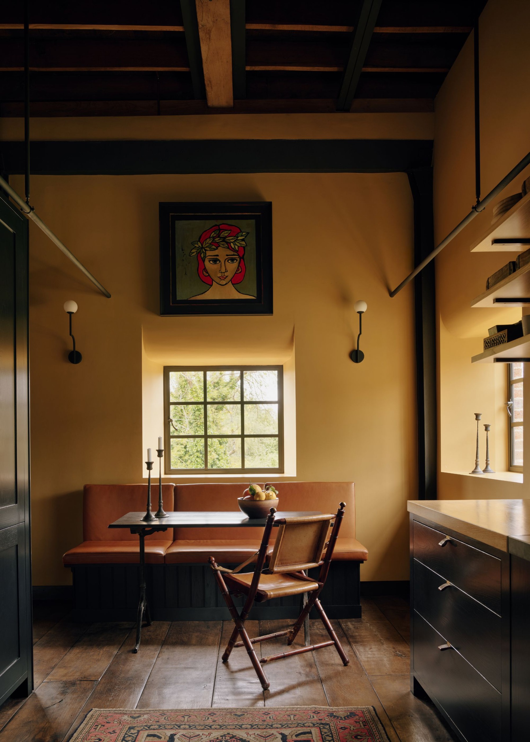 An industrial-style kitchen with a tall ceiling, wooden beams, and ochre walls. A small window with a tan bench, a dining table, and a chair. Dark wood flooring, black cabinets, and rustic decor.