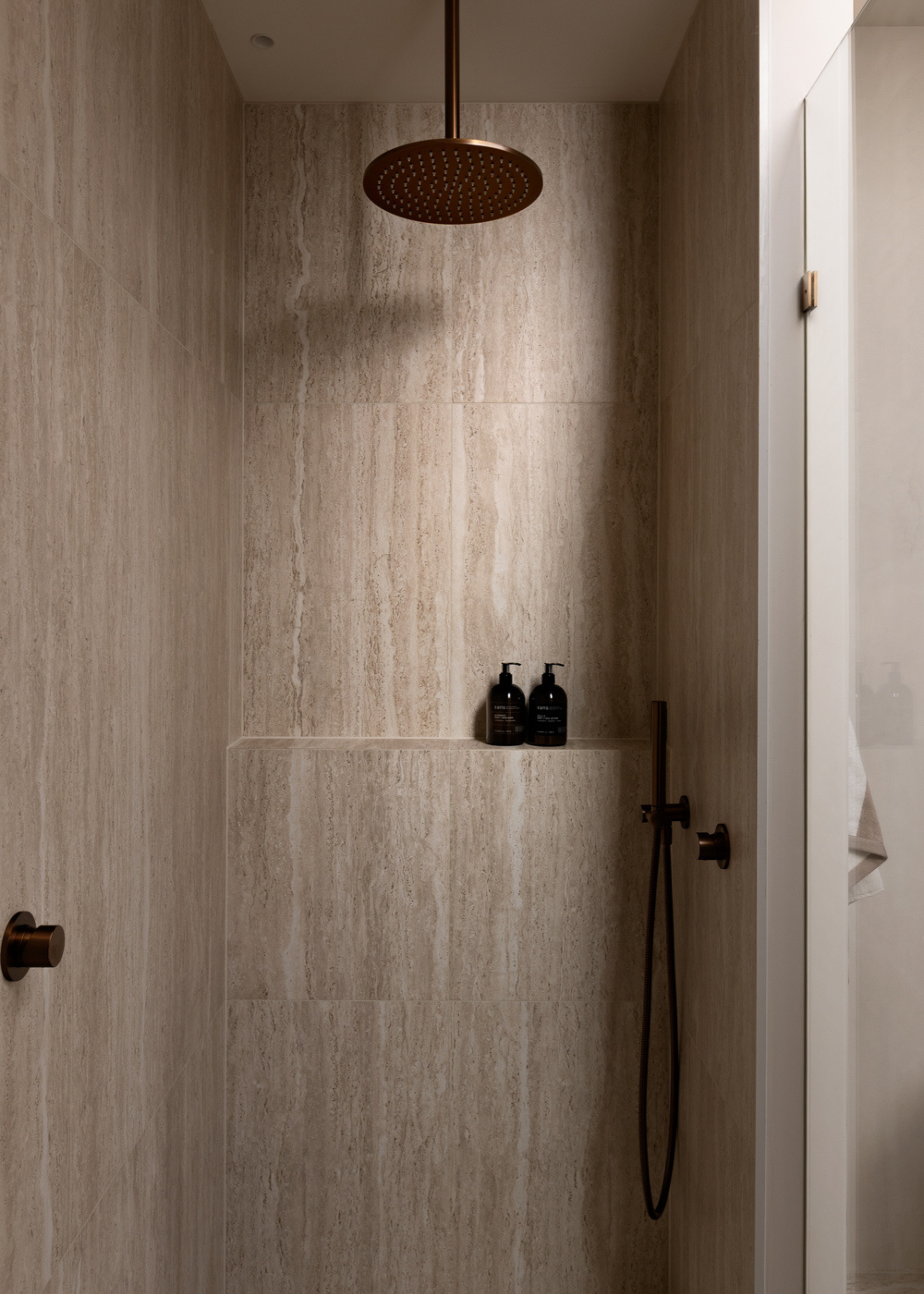 A beige bathroom with an overhead shower, a shelf for bath products, and black hardware