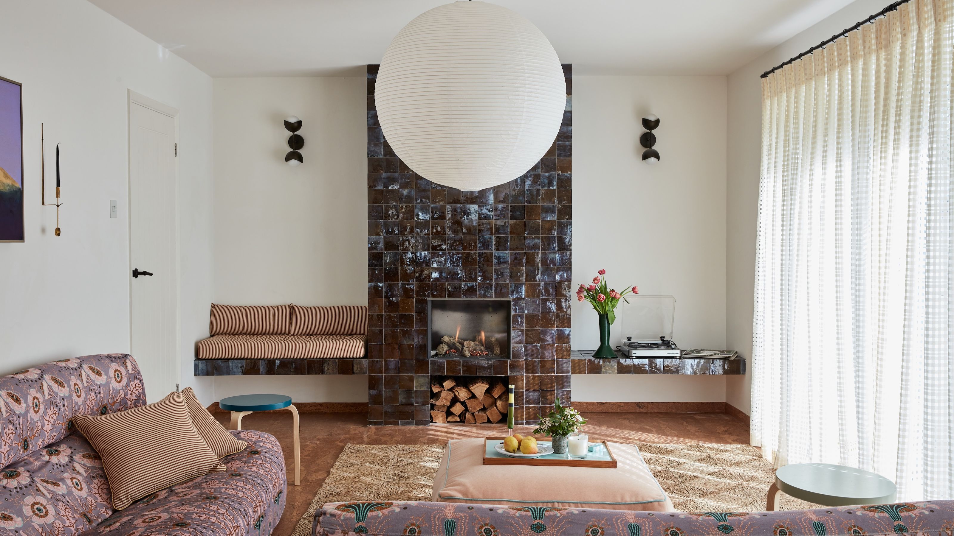 A modern take on a seventies style living room with a chimney breast tiled fully in brown tiles, a bench built in one side and a shelf the other. two sofas in and L-shape just infront with a coffee table and a big paper pendant lamp.