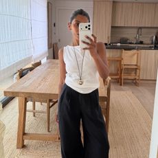 Woman wears white tank top, black linen balloon trousers, gold pendant necklace