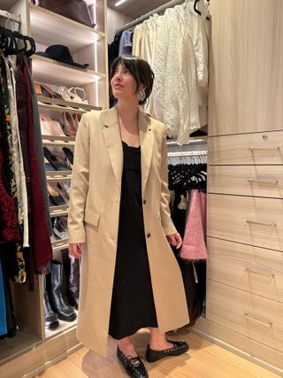 Kat stands in her closet wearing a beige coat, black dress, and black flats.