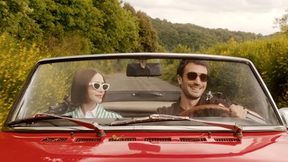 A young couple dressed in casual chic clothes sits with sunglasses on in a vintage red car while driving through the verdant countryside.
