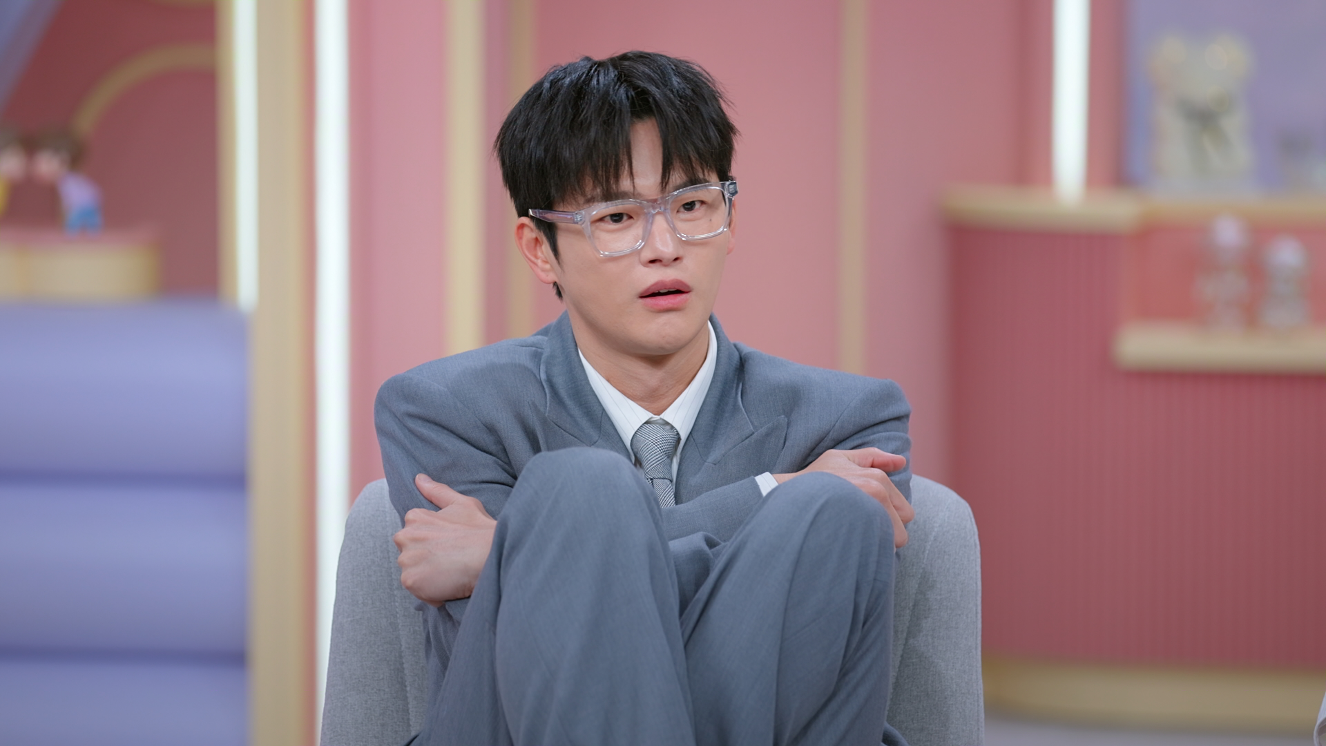 Co-host Seo In-guk hugs himself while sitting with his knees to his chest. He's perched in a gray chair on a light-pink film set, in a still from Netflix's 'Better Late Than Single.'