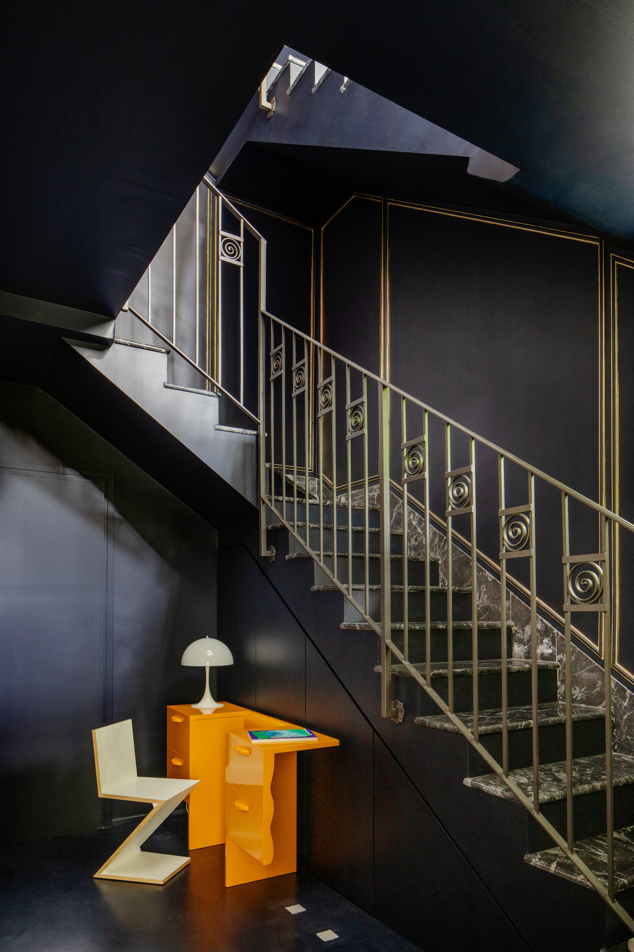 Black hallway with stairs adorned with gold balustrade, with bright orange desk and white zigzag chair underneath