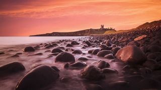 Rocky shoreline at sunset with a distant cliff and ancient castle ruins under an orange sky