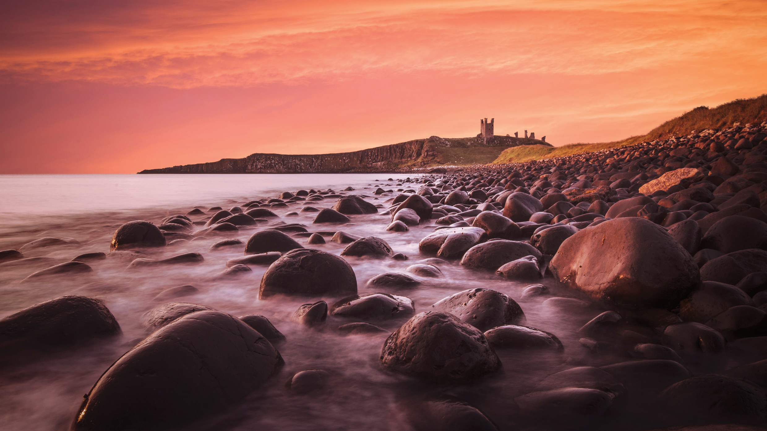 Rocky shoreline at sunset with a distant cliff and ancient castle ruins under an orange sky