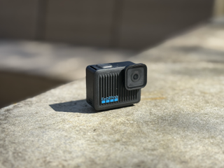 A small black camera with horizontal vents sits on a concrete slab in full sun