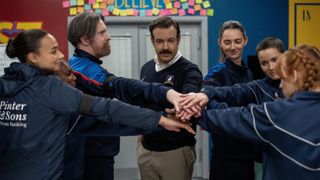 From left to right: Gemma, Shannon, Beard, Ted, Alice, Boots and another player putting their hands into the middle in Ted Lasso. 