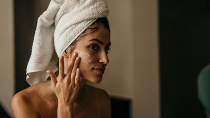 night-time skincare routine - A woman looking into the mirror with a towel on her head applying cream to her face 