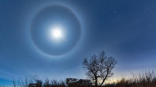 Clear night sky with a moon surrounded by a ring or halo with a house, tree and bracken in the foreground