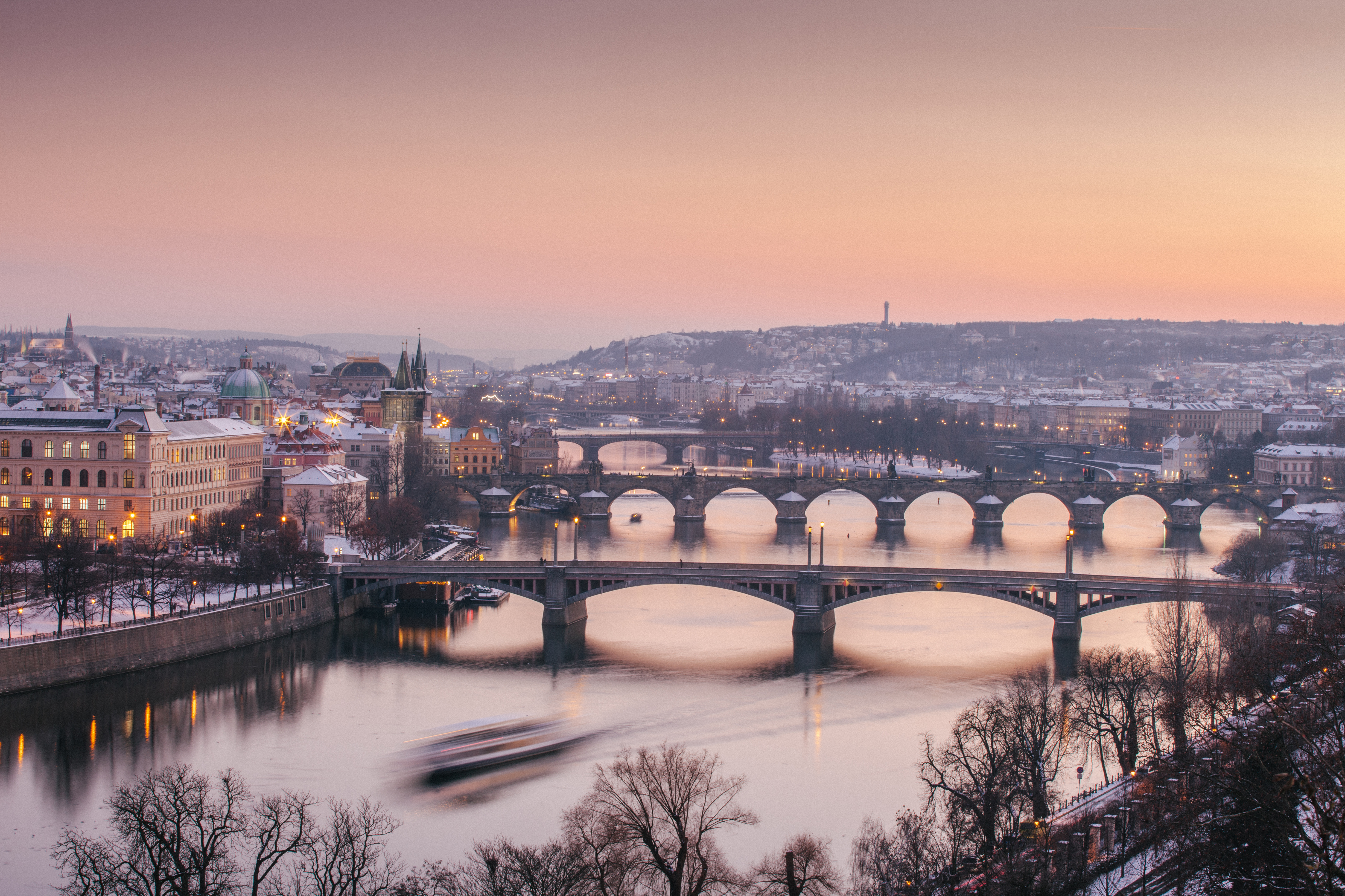 A sunset view over an ornate river city covered in a thin layer of snow, captured from above, shows beautifully preserved historical architecture, trams passing through its streets, and vegetation.