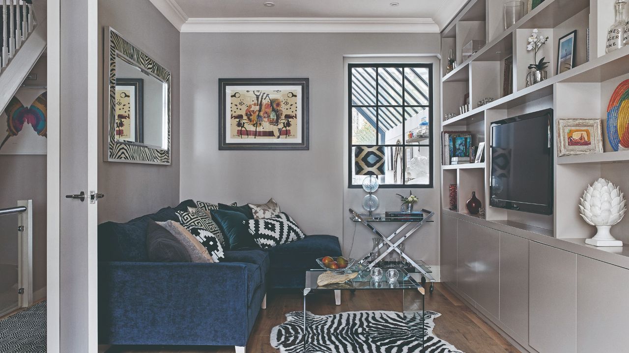 A grey-painted narrow living room with a navy velvet corner sofa and a media unit built alongside one wall housing the TV