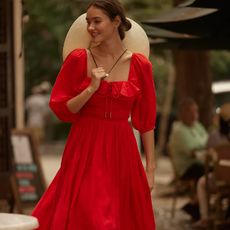 model wears red dress and sun hat while walking down a street