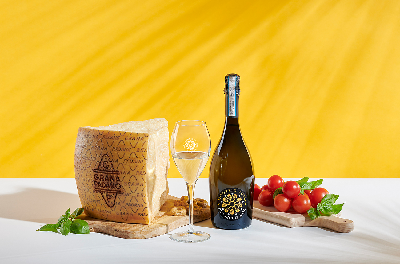 A large block of Grana Padano with a bottle of Prosecco and a little chopping board with tomatoes, on a white surface with a yellow background