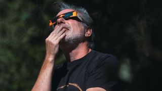 a man wearing eclipse glasses looks up at the sky and holds his had to his mouth.
