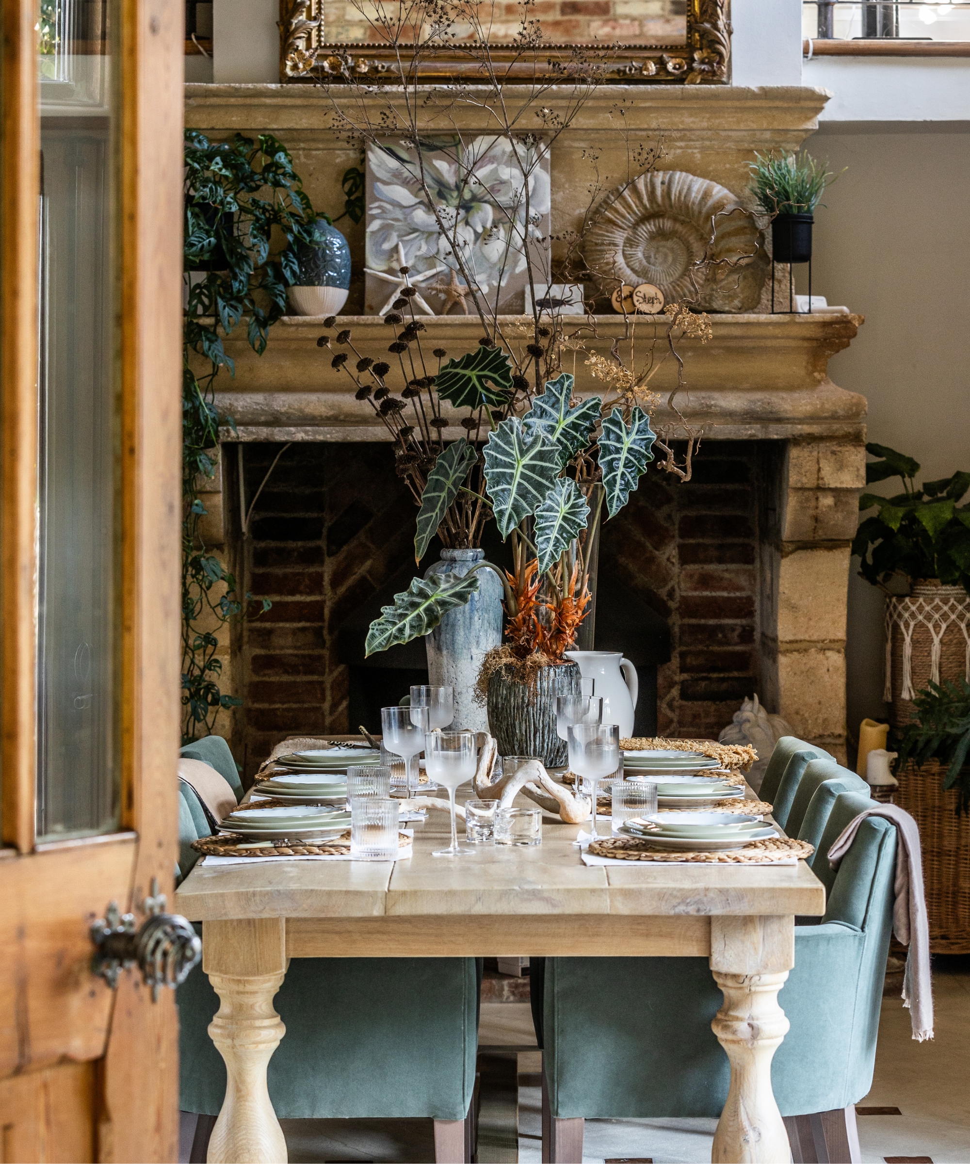 Set dining table before ornate stone fireplace, green velvet chairs, ammonite fossil, alocasia leaves.