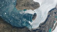 A satellite photo of a fjord with water covered in tiny icebergs and an wave of water arcing across the surface