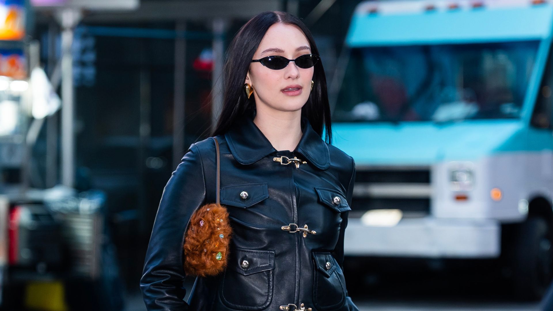 Lola Tung stands on the road in New York wearing a leather toggle jacket with low-rise jeans, cow print trainers and a furry bag.
