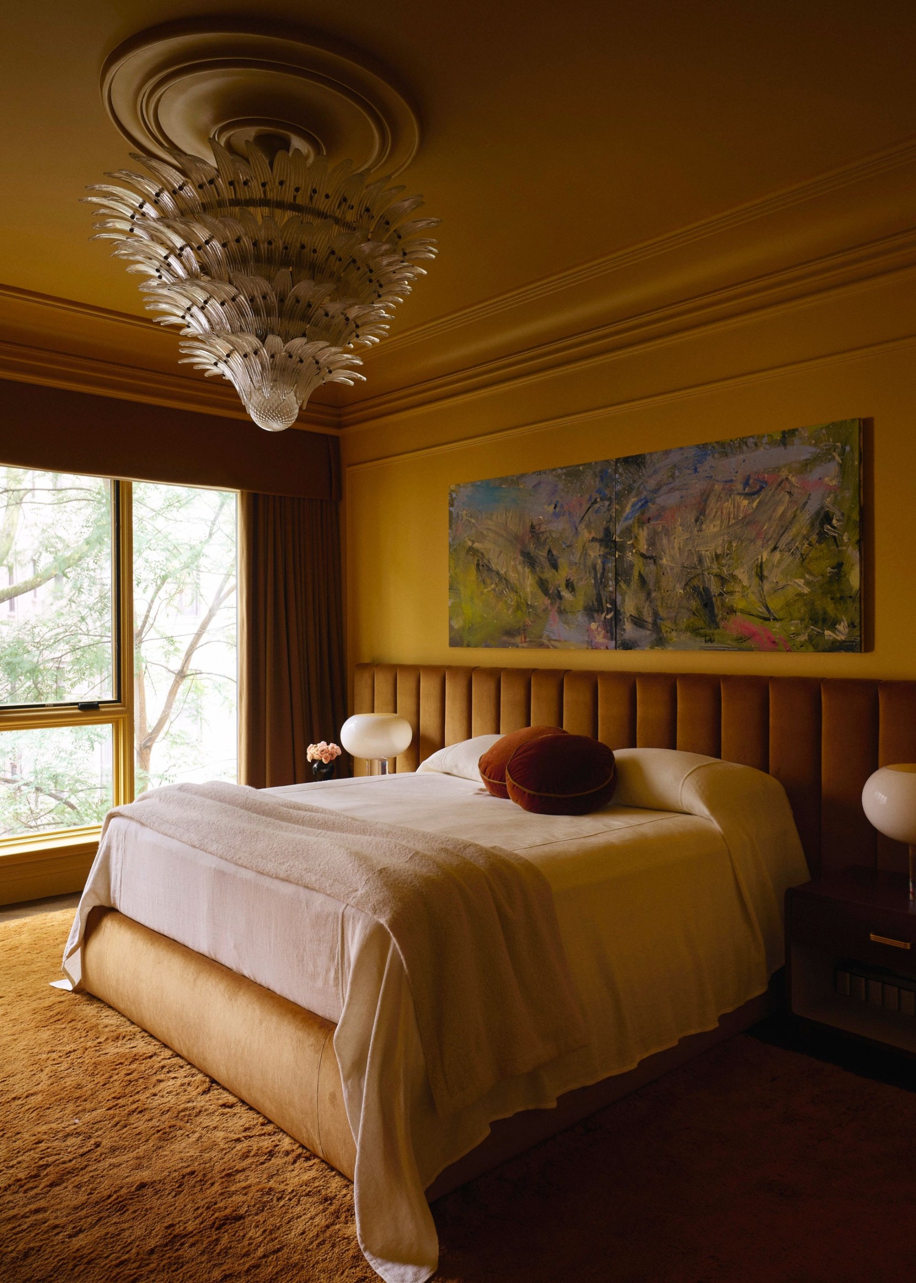 A modern bedroom with ochre color-drenched walls with decorative moldings, a large bed with neutral bedding, and a caramel-brown elongated headboard. A warm brown textured rug and large windows.