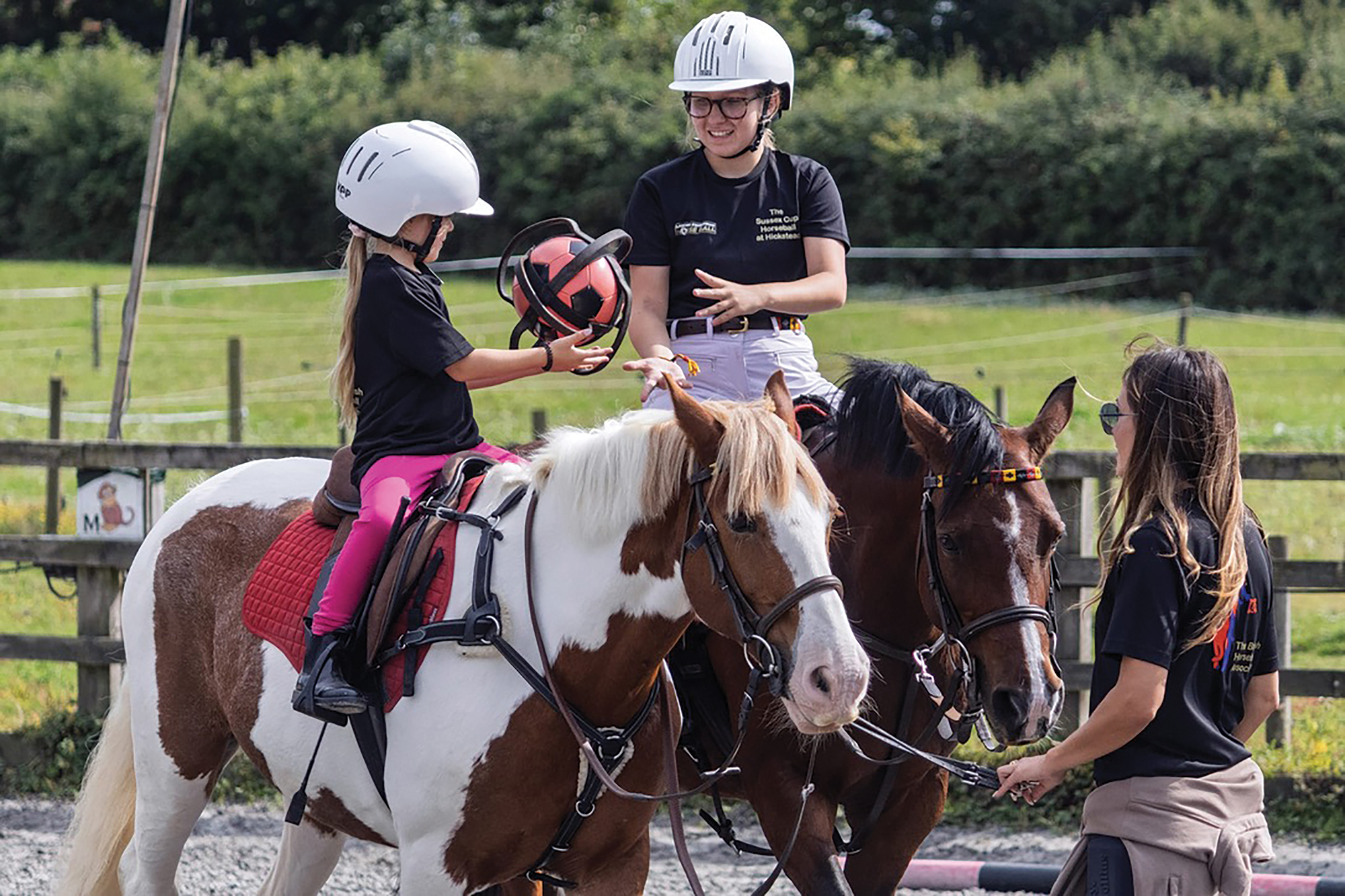 Two young riders toss a horseball from one to the other while a lady holds both ponies. One pony is chestnut and white. The other pony is bay.