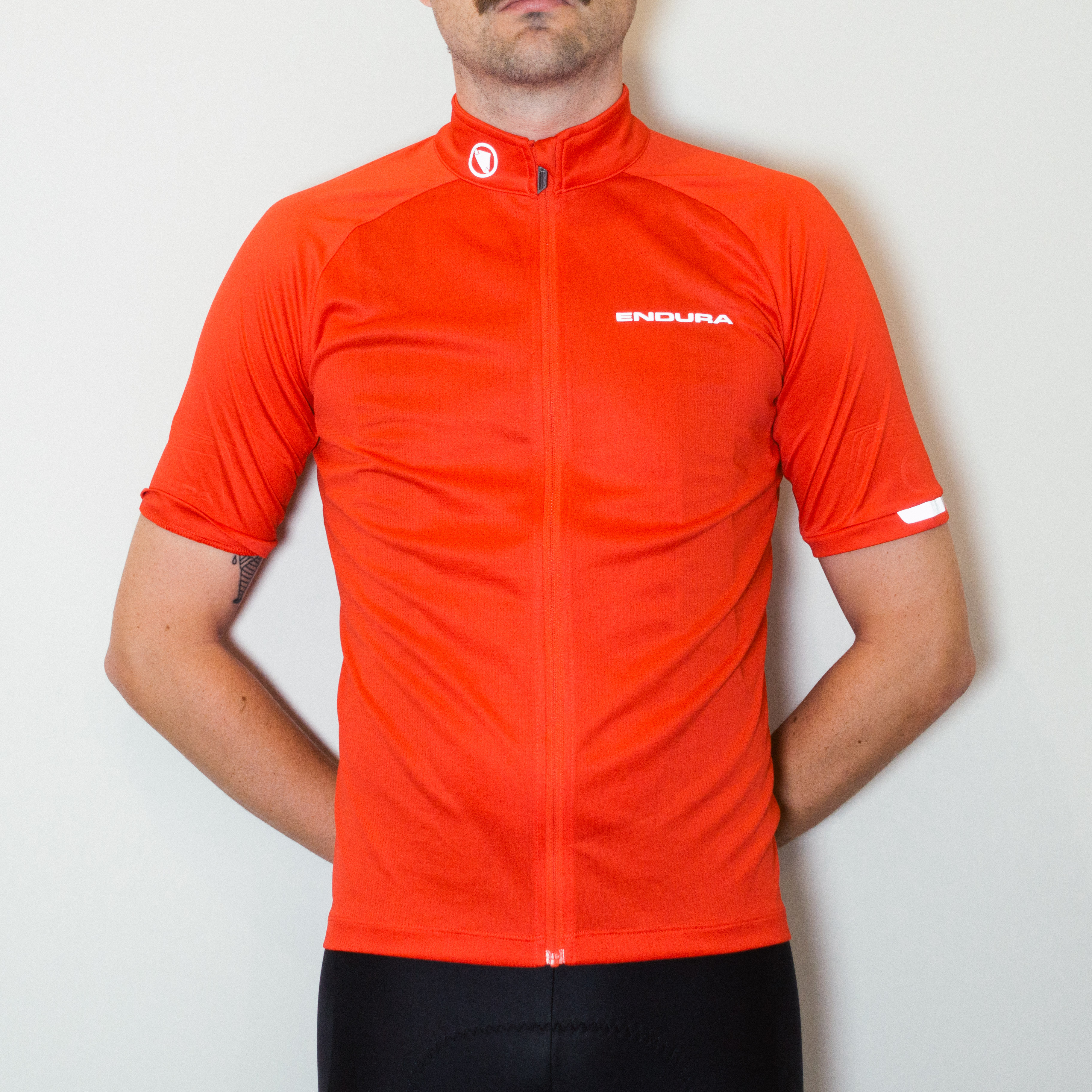 A white man in a red cycling jersey and black cycling shorts stands against a white wall