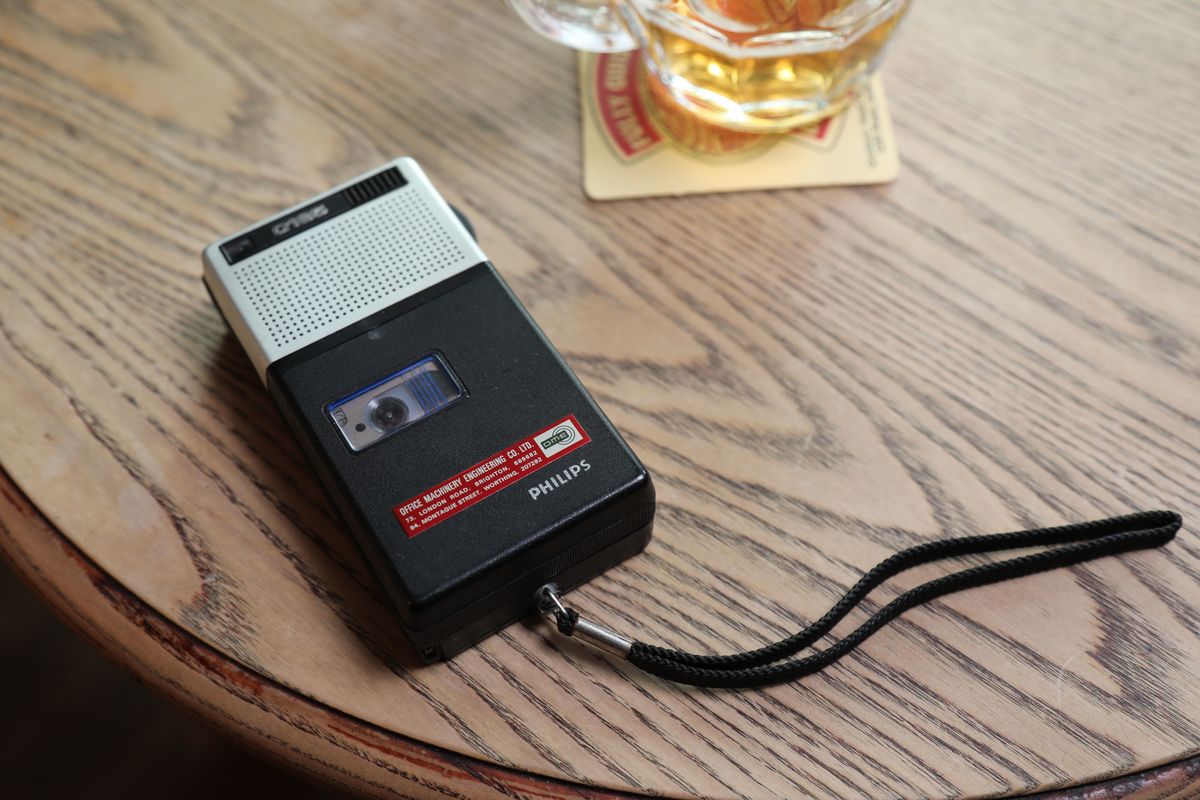 AIDS: The Unheard Tapes - a picture of a Philips dictaphone recording on an audio cassette, sitting on the edge of a wooden table next to a pint tankard of beer