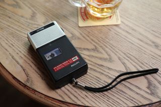 AIDS: The Unheard Tapes - a picture of a Philips dictaphone recording on an audio cassette, sitting on the edge of a wooden table next to a pint tankard of beer