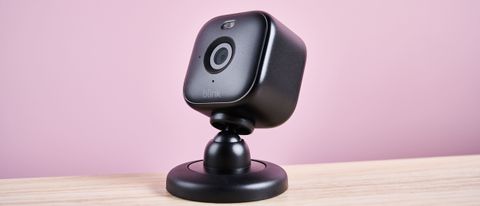 A black-colored Blink Mini 2 sits on a light wooden surface against a pink background. The camera is angled up towards the top left corner of the image.