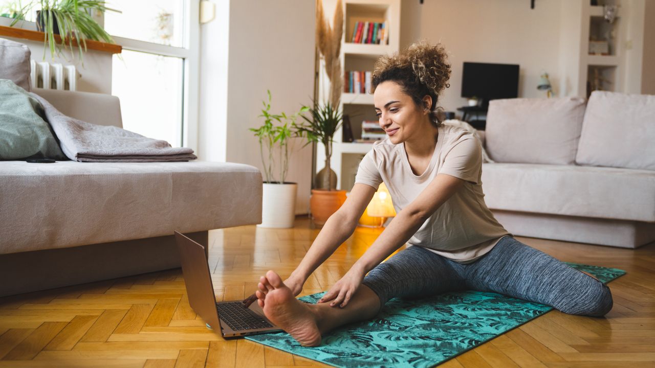 A woman sits on a yoga mat in her living room, with one leg stretched in front of her and the other bent to the side. Her fingertips reach towards her toes on her extended leg. Behind her we see couches, shelving and plants.