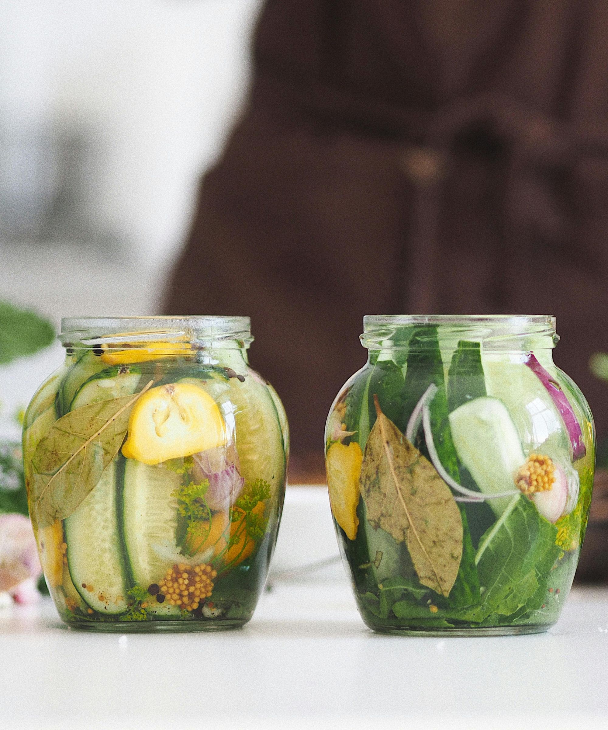 Two large mason jars filled with sliced cucumbers, sliced lemons and salad leaves, sitting on a white table in front of a figure wearing a brown apron