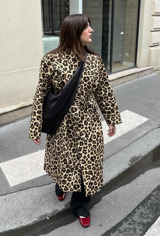 The leopard print coat trend is shown on a woman standing outside wearing a long leopard coat over black trousers styled with red ballet flats and a black sling bag