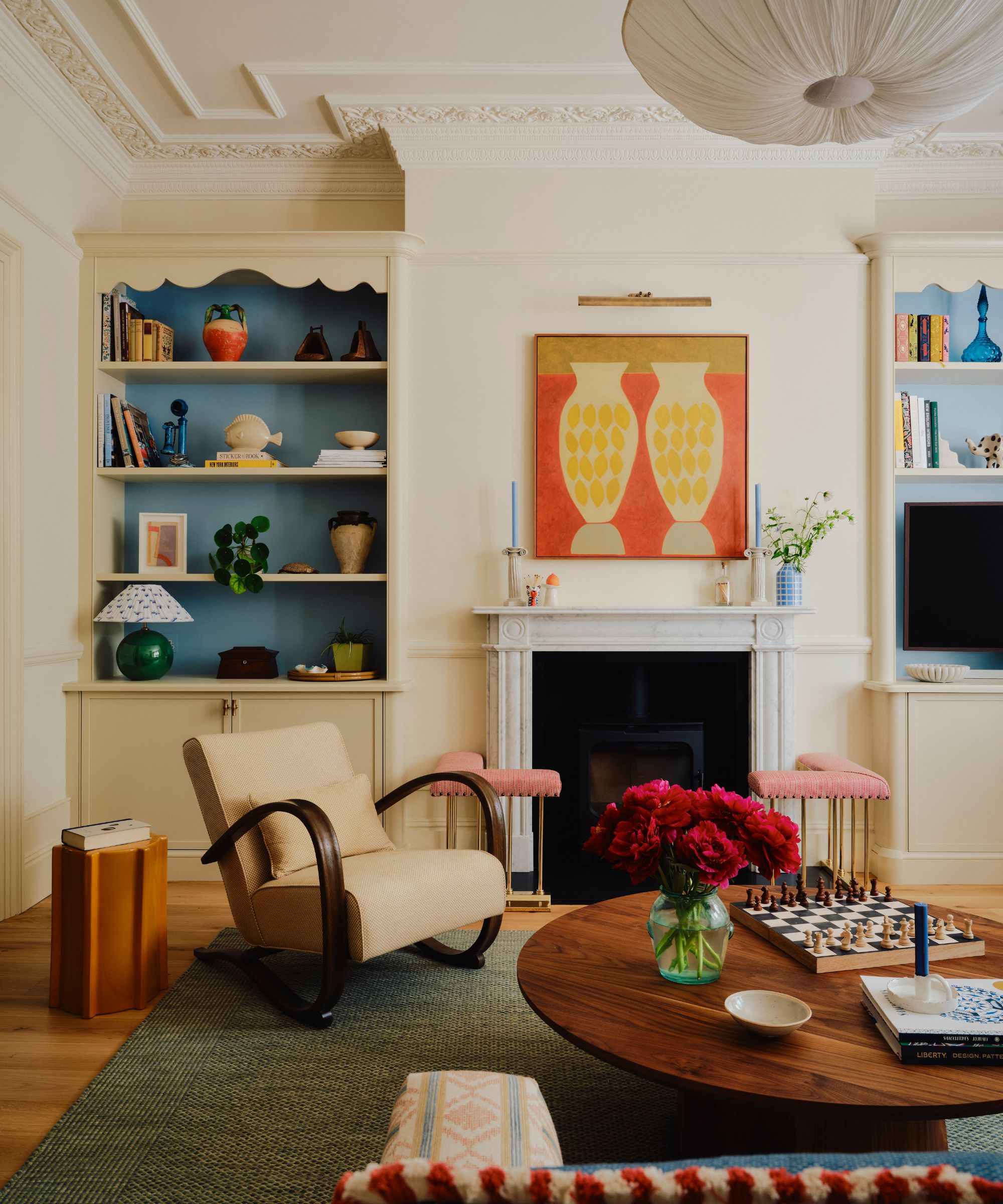 A large living room with cream walls, crown moulding, a large marble fireplace with a pink hearth, a low, round wooden coffee table, a mid-century armchair, and blue-painted shelving alcoves.