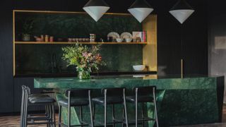 A kitchen island in green marble stone with gold open shelving behind and white pointed pendant lighting above