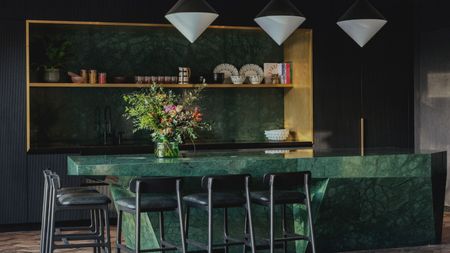 A kitchen island in green marble stone with gold open shelving behind and white pointed pendant lighting above