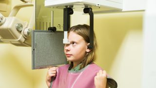 Caucasian blonde girl stands inside a panoramic dental x-ray machine at the orthodontist. She wears a ping lead apron over her shirt.