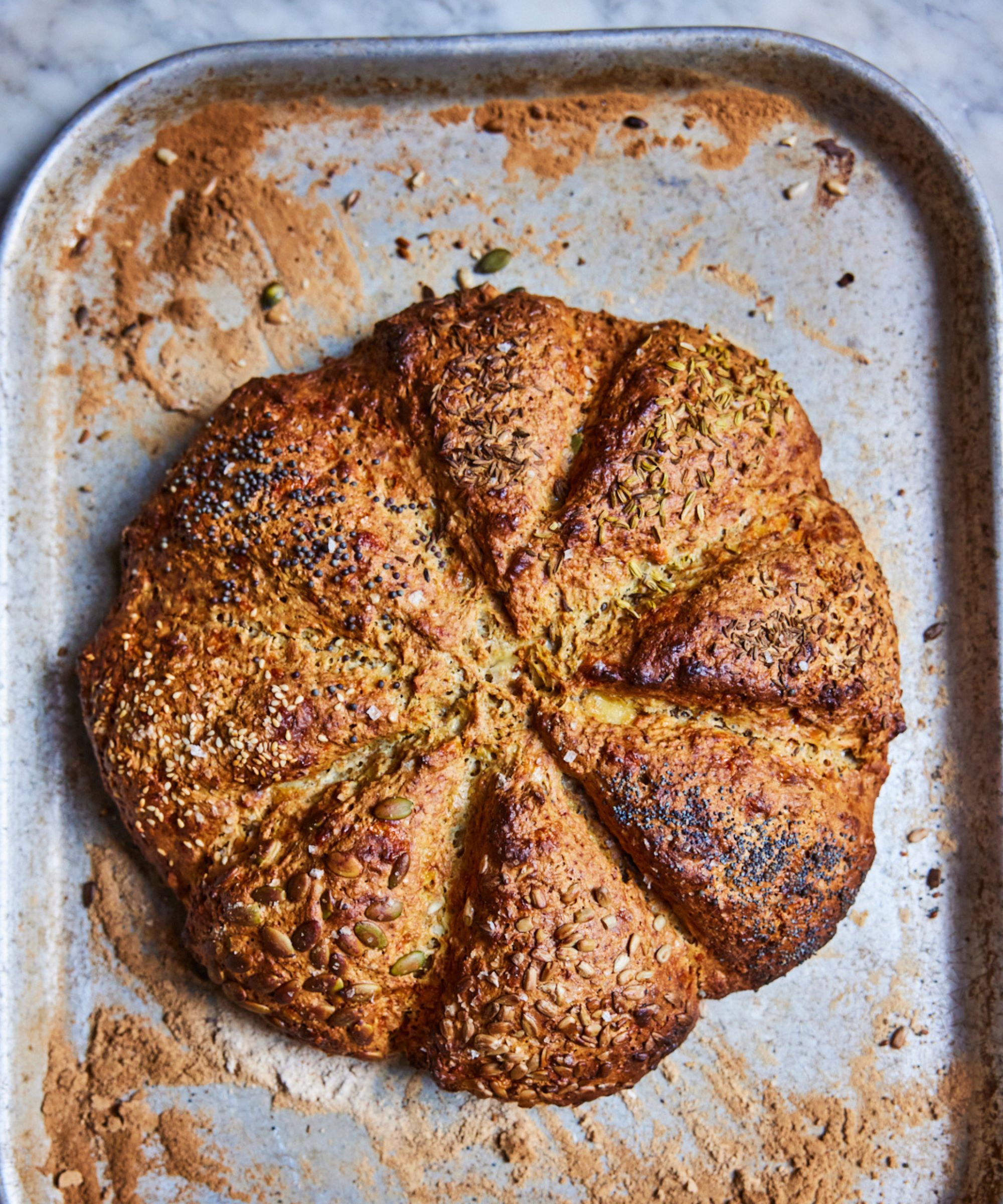 Round loaf of seeded bread on a rustic baking tray covered in a dusting of flour and seeds