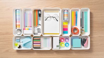 Neatly organized stationery in white trays on wooden desk viewed from above
